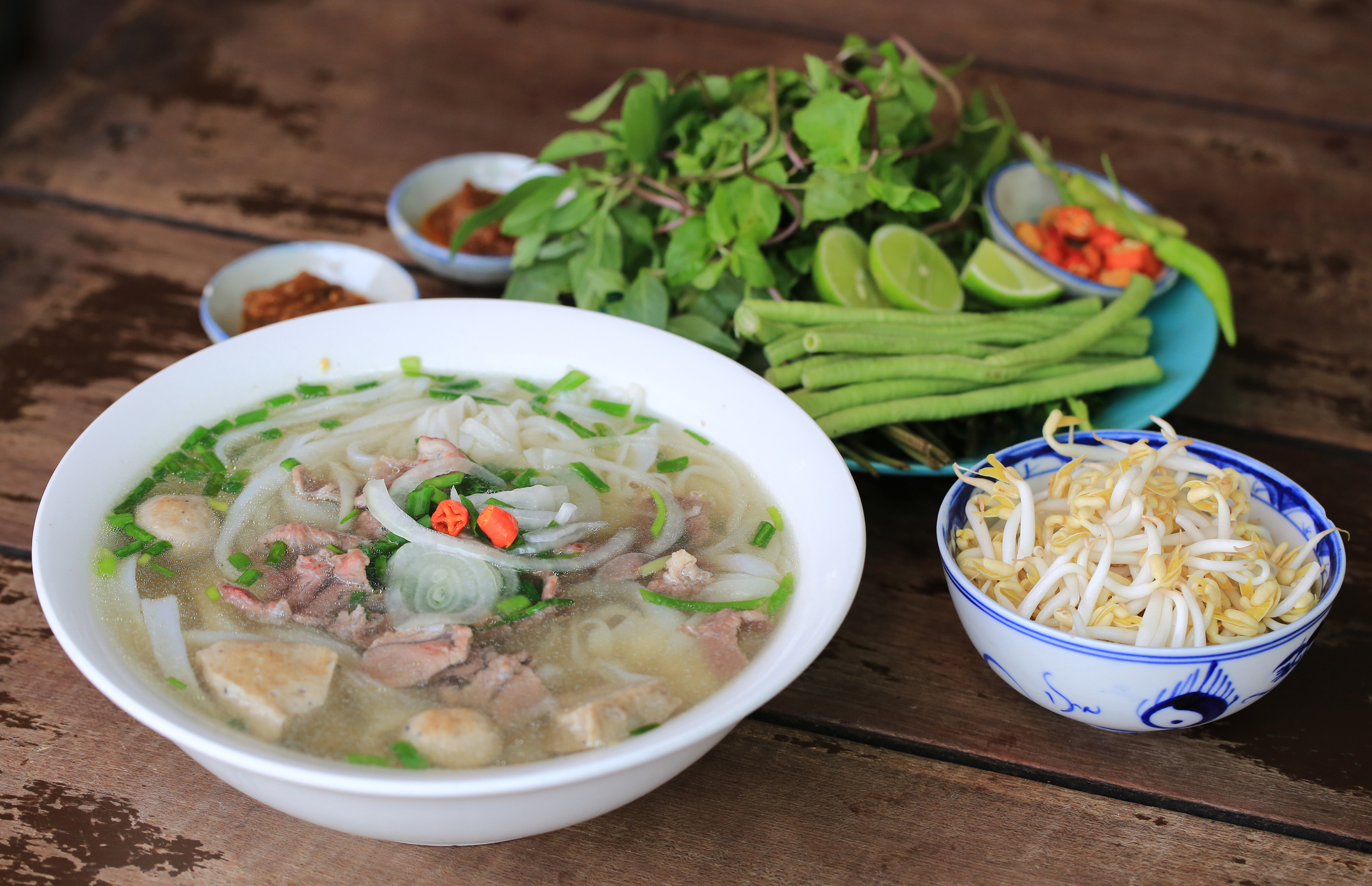 A bowl of Pho accompanied by a plate of green vegetables and a small bowl of bean sprouts
